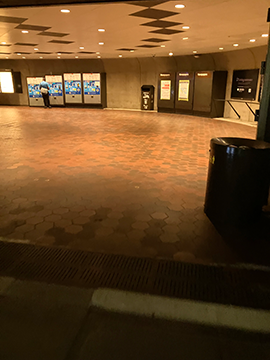 A picture of a pathway in a metro station. In the foreground to the right there is a trash can. In the background there is a wall with fare vending machines slightly to the right of center as well as to the left. In the far right background there is the ending of a railing. A picture of a pathway in a metro station. In the foreground to the right there is a trash can. In the background there is a wall with fare vending machines slightly to the right of center as well as to the left. In the far right background there is the ending of a railing.